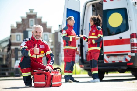 Photo for Joyful paramedic seated on his haunches before the medical bag making the thumbs-up sign while his colleagues talking - Royalty Free Image