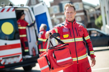 Téléchargez les photos : Souriant heureux ambulancier caucasien dans l'uniforme rouge donnant son sac EMS à un collègue - en image libre de droit