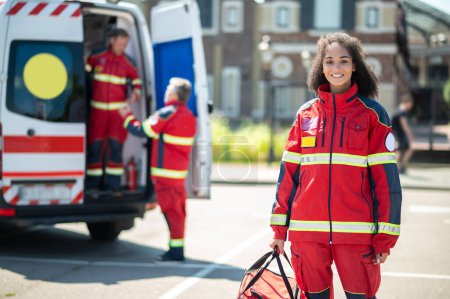 Téléchargez les photos : Jeune ambulancière souriante avec un sac médical debout à l'extérieur et attendant ses collègues - en image libre de droit