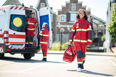 Photo for Smiling joyous female paramedic with an EMS bag in the hand walking along the street - Royalty Free Image