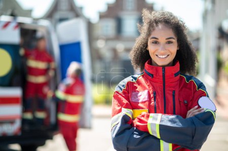 Foto de Retrato en la cintura de una doctora sonriente y confiada en la ambulancia parada al aire libre con los brazos cruzados y mirando hacia el futuro - Imagen libre de derechos
