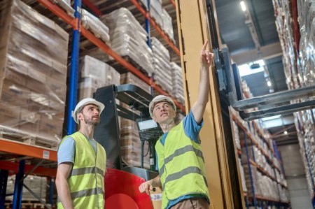 Smiling middle-aged stockhouse worker standing beside his young colleague and pointing upward at pallet racks
