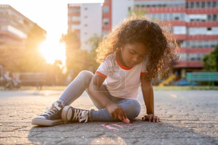 Photo for Concentrated little girl sitting on the paved ground and drawing with a piece of colored chalk - Royalty Free Image