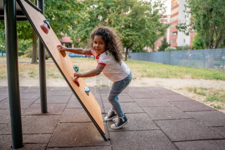 Photo for Sporty little African American girl posing for the camera while scaling the climbing wall on the playground - Royalty Free Image