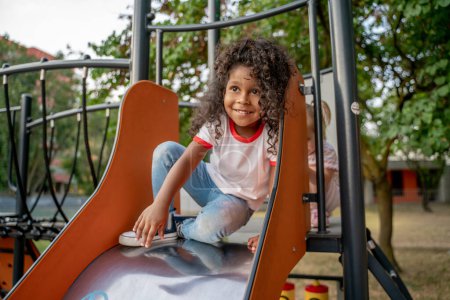 Photo for Smiling cute little African American girl and her friend sitting at the top of the stainless steel slide - Royalty Free Image