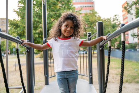 Photo for Smiling pleased curly-haired little girl leaning on the children slide handrails and looking before her - Royalty Free Image