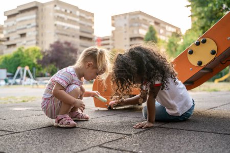 Photo for Serious focused cute little blonde girl and her curly-haired friend drawing with colored chalks on the ground - Royalty Free Image