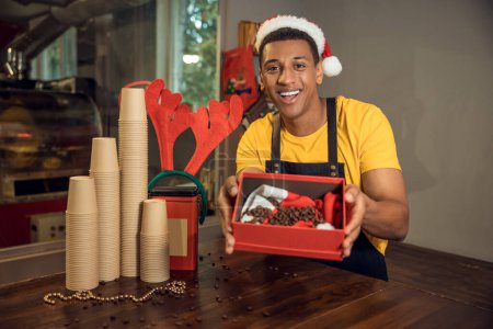 Photo for Smiling cheerful barista in the Santa hat showing a gift box with roasted coffee beans in front of the camera - Royalty Free Image