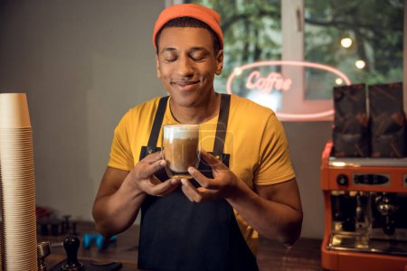 Téléchargez les photos : Portrait taille haute d'un jeune barista afro-américain souriant jouissant de l'arôme du cappuccino - en image libre de droit