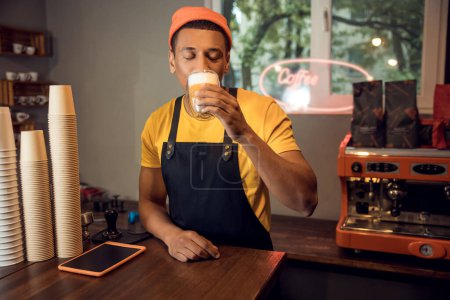 Photo for Waist-up portrait of an African American barista drinking cappuccino from the glass cup at the table - Royalty Free Image