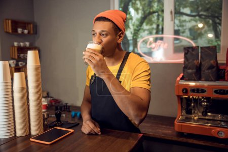 Téléchargez les photos : Portrait taille haute d'un barista buvant du café dans la tasse en verre à la table et regardant au loin - en image libre de droit