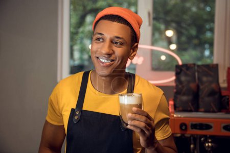 Photo for Portrait of a cheerful cafe worker holding a glass of cappuccino in the hand and looking away - Royalty Free Image