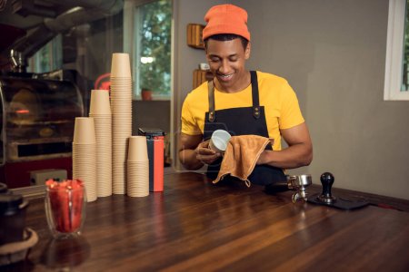 Photo for Waist-up portrait of a smiling cafe worker standing at the table while wiping the cup with a microfiber towel - Royalty Free Image