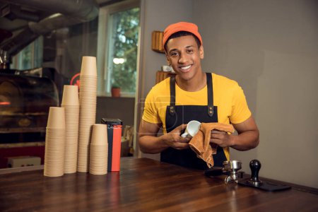 Foto de Retrato en la cintura de un trabajador de café sonriente y complacido limpiando una taza con una toalla de microfibra - Imagen libre de derechos