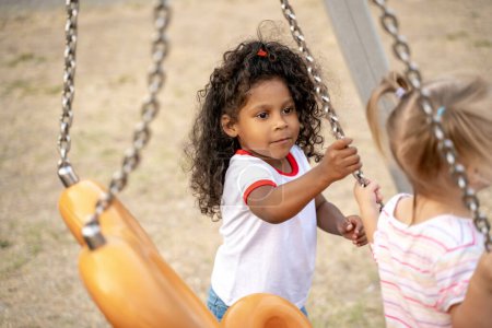 Photo for Strong cute little child with curly hair pushing a blonde girl on the playground swing - Royalty Free Image