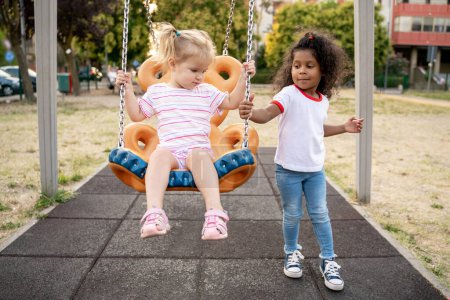 Photo for Strong kid standing on the playground and pushing a blonde little girl on the swing - Royalty Free Image