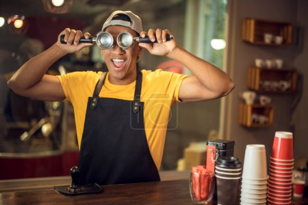Photo for Waist-up portrait of a cheerful barista covering his eyes with a pair of portafilter baskets - Royalty Free Image