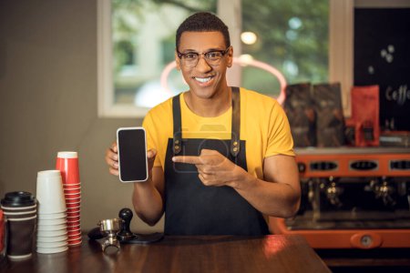 Foto de Retrato en la cintura de un alegre camarero apuntando con el dedo índice al smartphone de la mano - Imagen libre de derechos