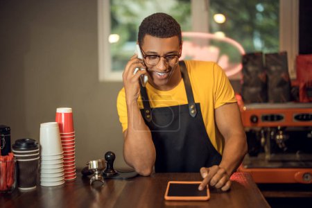 Photo for Waist-up portrait of a joyous barista seated at the table using his tablet computer during the phone conversation - Royalty Free Image
