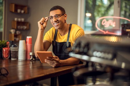 Téléchargez les photos : Souriant heureux jeune barista assis à la table avec une tablette dans la main - en image libre de droit