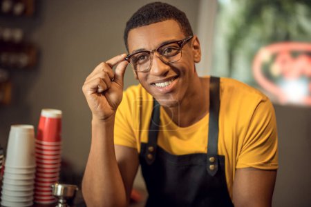 Photo for Portrait of a smiling happy coffee shop worker touching the rim of his spectacles and looking into the distance - Royalty Free Image