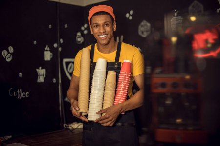 Foto de Sonriendo satisfecho trabajador de la cafetería en el delantal sosteniendo una pila de vasos de papel invertido desechables en sus manos - Imagen libre de derechos