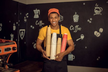 Photo for Smiling contented African American barista holding stacked disposable inverted paper coffee cups in his hands - Royalty Free Image