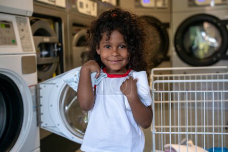 Foto de Retrato en la cintura de una niña sonriente y complacida con ropa en las manos posando para la cámara en la lavandería - Imagen libre de derechos