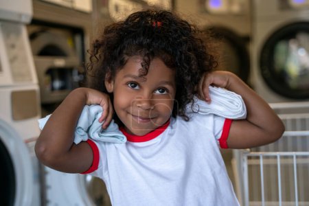 Photo for Cute smiling happy kid with clean clothes in her hands posing for the camera in a laundry room - Royalty Free Image