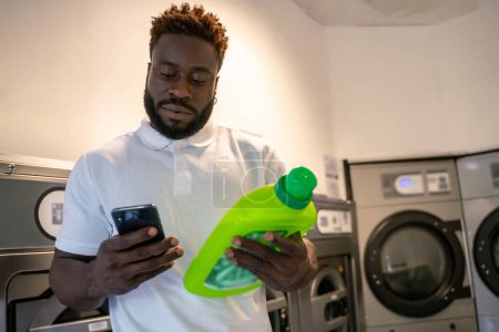 Foto de Retrato en la cintura de un joven serio sosteniendo una botella de detergente mientras mira su teléfono celular - Imagen libre de derechos