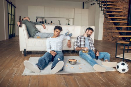 Photo for Tipsy young man with a bottle of beer sitting on the floor while his pals dozing off - Royalty Free Image