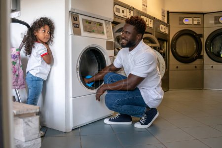 Téléchargez les photos : Homme souriant assis sur ses hanches plaçant la lessive dans une machine à laver tout en regardant son enfant - en image libre de droit