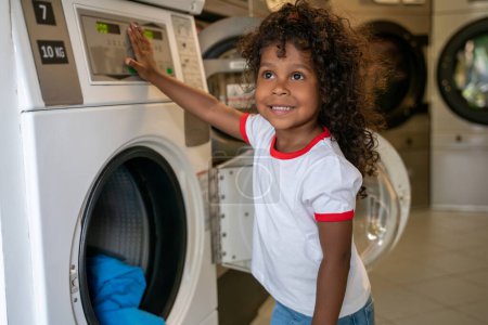 Photo for Smiling pleased little girl leaning on the washing machine control panel and looking into the distance - Royalty Free Image