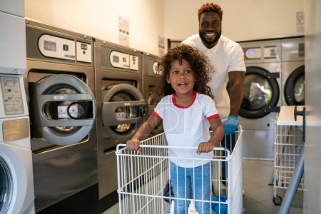 Photo for Cheerful young African American man wheeling his happy cute little daughter in the metal laundry cart - Royalty Free Image