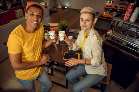 Photo for Smiling young coffee shop worker standing beside his joyous female colleague seated at the table - Royalty Free Image