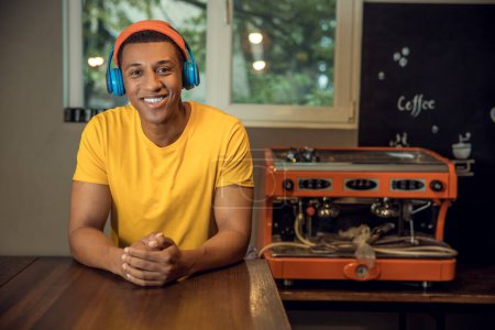 Foto de Retrato en la cintura de un joven trabajador de la cafetería alegre en los auriculares inalámbricos apoyados en la mesa y mirando hacia el futuro - Imagen libre de derechos