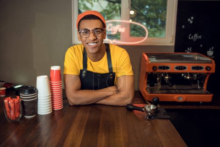 Photo for Waist-up portrait of a joyous barista in the eyeglasses leaning on the table and looking before him - Royalty Free Image