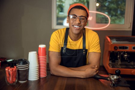 Foto de Retrato en la cintura de un trabajador del café alegre en los espectáculos apoyados en la mesa y mirando hacia el futuro - Imagen libre de derechos
