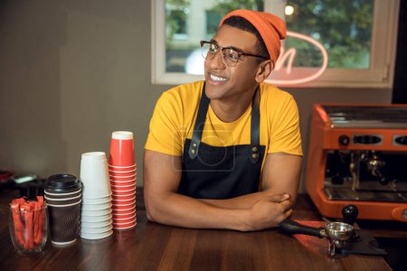 Photo for Waist-up portrait of a pleased coffee worker in the eyewear leaning on the table and looking away - Royalty Free Image