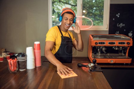 Foto de Retrato en la cintura de un alegre camarero en los auriculares inalámbricos limpiando la mesa con un paño - Imagen libre de derechos