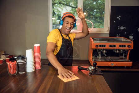 Foto de Retrato en la cintura de un camarero feliz en los auriculares limpiando la mesa de la cafetería con música - Imagen libre de derechos