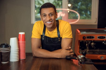 Photo for Waist-up portrait of a joyful barista leaning on the cafe table and looking in front of him - Royalty Free Image