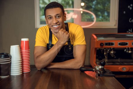 Foto de Retrato en la cintura de un trabajador de una cafetería alegre apoyado en la mesa y mirando delante de él - Imagen libre de derechos