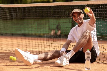 Foto de Pista de tenis. Joven jugador de tenis guapo sentado en la cancha de tenis con una pelota de tenis en las manos - Imagen libre de derechos
