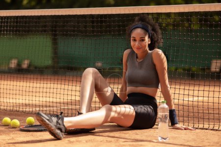 Téléchargez les photos : Jolie fille au court de tennis. Jolie fille aux cheveux bruns assis sur le court de tennis et regardant satisfait - en image libre de droit