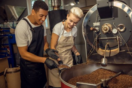 Téléchargez les photos : Maître rôti souriant montrant une cuillère en métal pleine de grains de café torréfiés à son collègue heureux - en image libre de droit