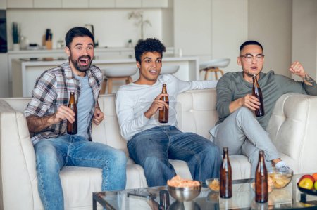 Foto de Tres hombres con botellas de cerveza sentados en el sofá y viendo un partido de fútbol - Imagen libre de derechos