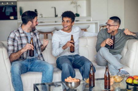 Photo for Cheerful guy with a bottle of beer sitting on the sofa in the company of his friends - Royalty Free Image