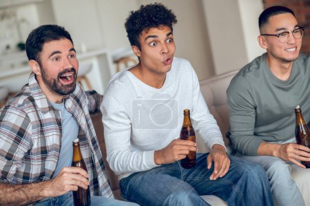 Foto de Tres jóvenes sorprendidos sentados en el sofá con botellas de cerveza y viendo algo - Imagen libre de derechos