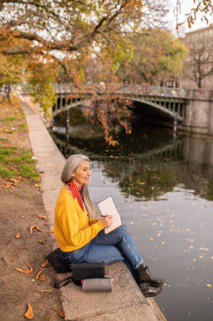 Foto de Humor creativo. Mujer sentada en la orilla del río y tomando notas - Imagen libre de derechos